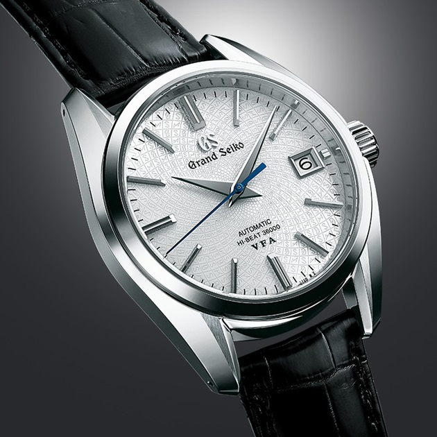 A close-up of a Grand Seiko watch angled to the left in the middle of a gray background. The watch features a textured white dial, stainless steel case, and black leather strap.