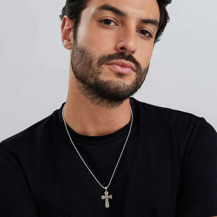 A man with dark hair and a beard wears a black shirt and a cross amulet necklace around his neck. The necklace features yellow caviar beading, striped detailing, and a ball chain.