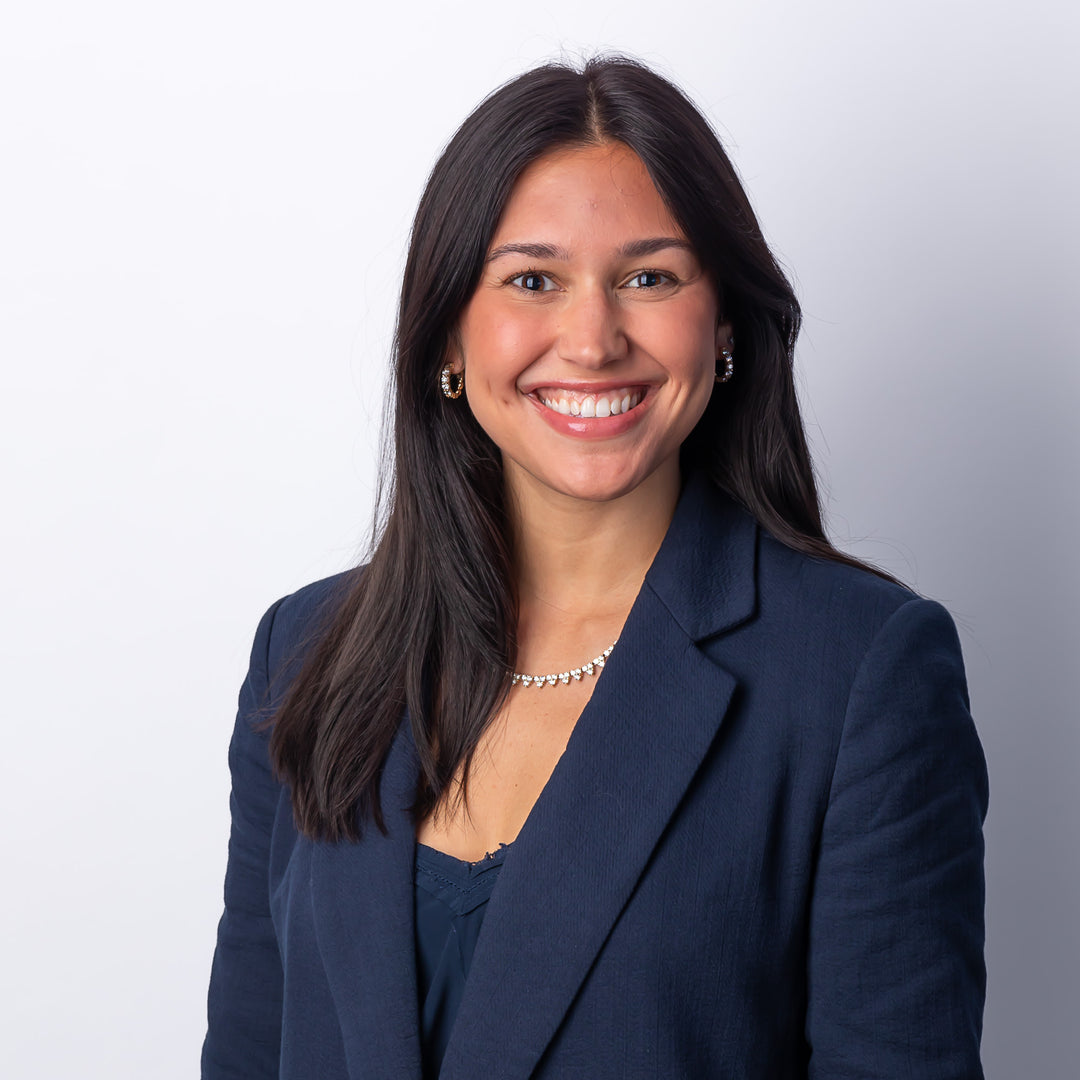 A headshot of a woman smiling, wearing a navy blouse and blazer with hoop earrings and a diamond necklace.