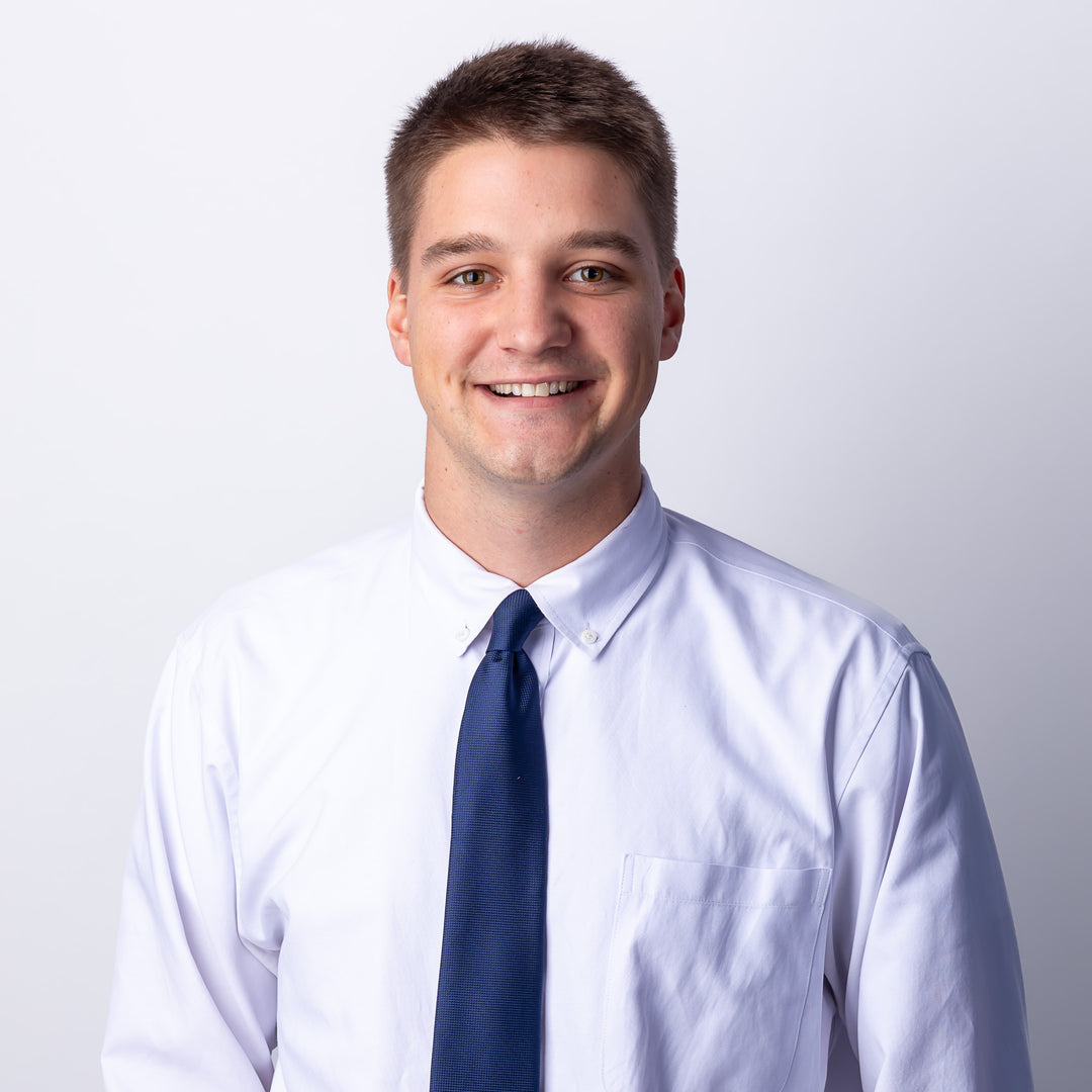 A headshot of a man with short brown hair smiling, wearing a white button-down and blue tie.