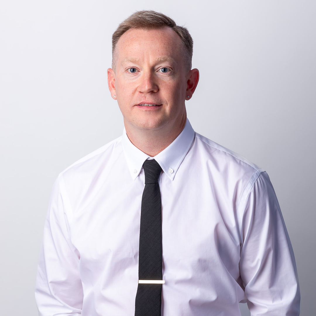 A headshot of a man with short hair smiling, wearing a button-down and a black tie.