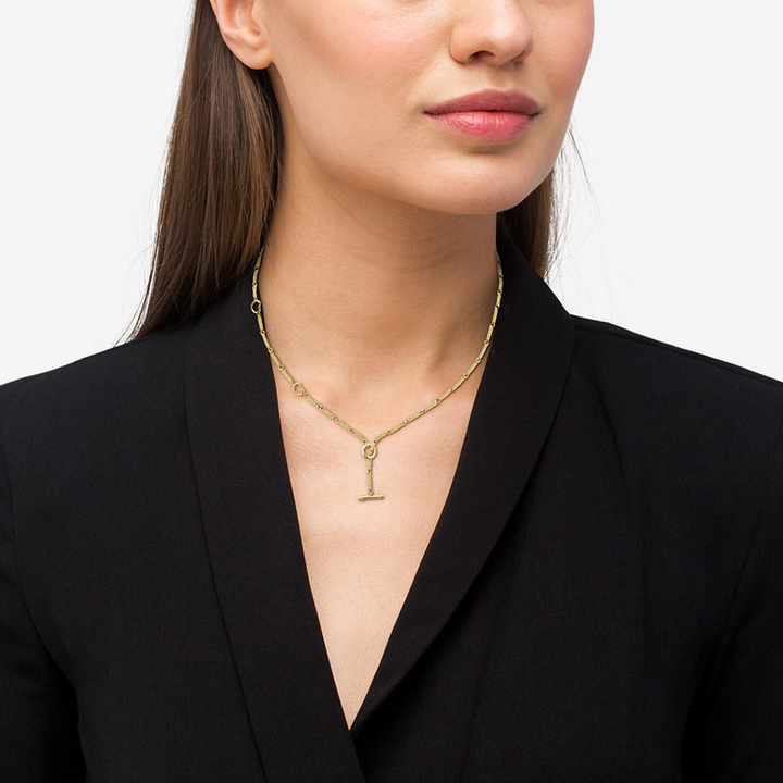 A close-up of a woman wearing a yellow gold necklace and black blouse. The necklace features a toggle design with heart-shaped stations.