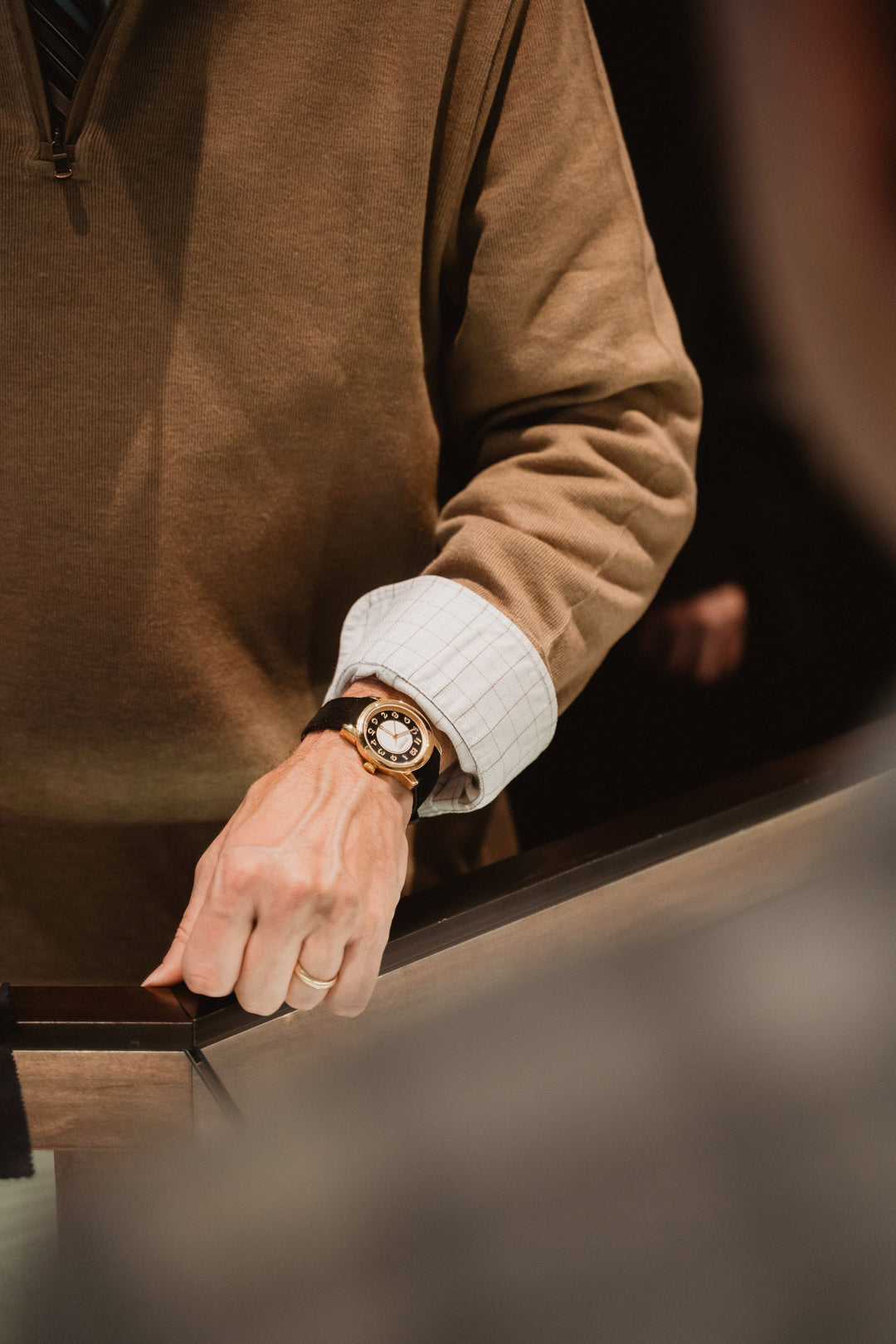 A close-up of a man wearing a brown sweater and a watch around his wrist. The watch features a black dial, a yellow gold bezel and case, and a black fabric strap.