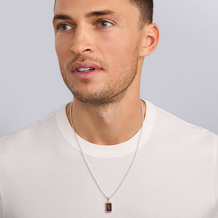 A close-up of a man wearing a white shirt and an amulet around his neck. The amulet features a brown textured center stone, caviar beaded edging, LAGOS engraved at the bottom, and a ball chain.