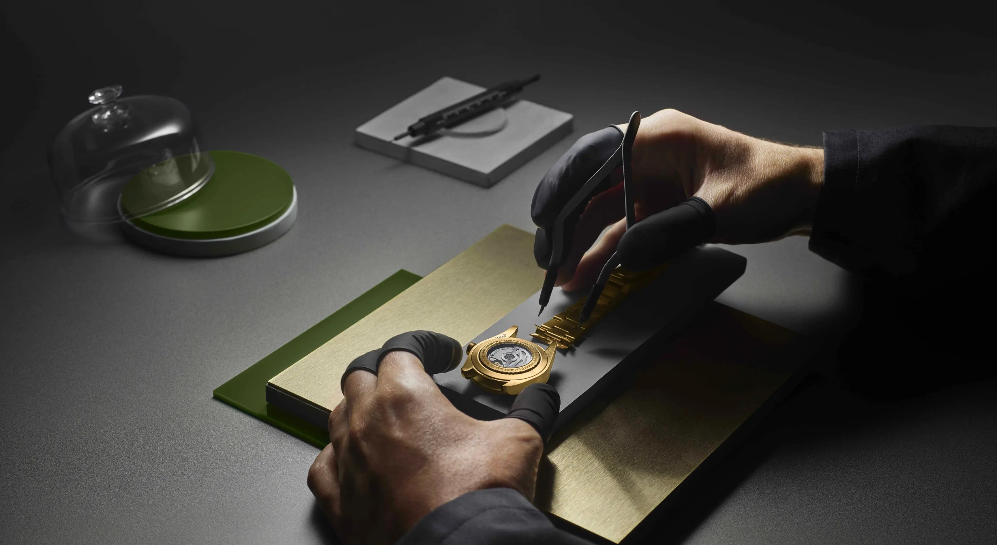 A person wearing black fingerless gloves works on a watch's dial with a yellow gold coating, using tools on a dark surface. A person wearing black fingerless gloves works on a watch's dial with a yellow gold coating, using tools on a dark surface.