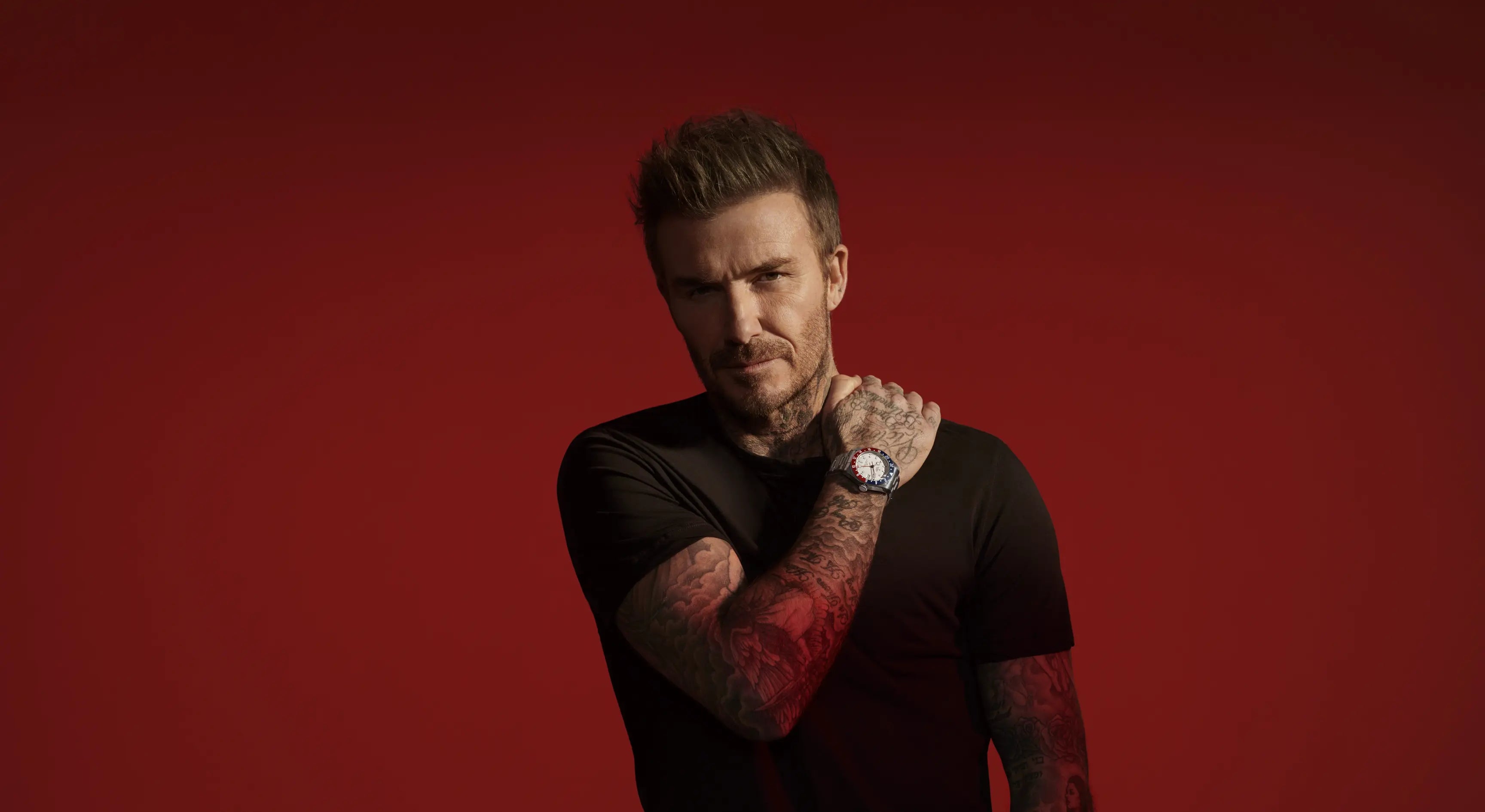 A headshot of David Beckham wearing a black t shirt, Tudor watch with a black and blue bezel, white dial, and stainless steel steep, standing against a red background. A headshot of David Beckham wearing a black t shirt, Tudor watch with a black and blue bezel, white dial, and stainless steel steep, standing against a red background.