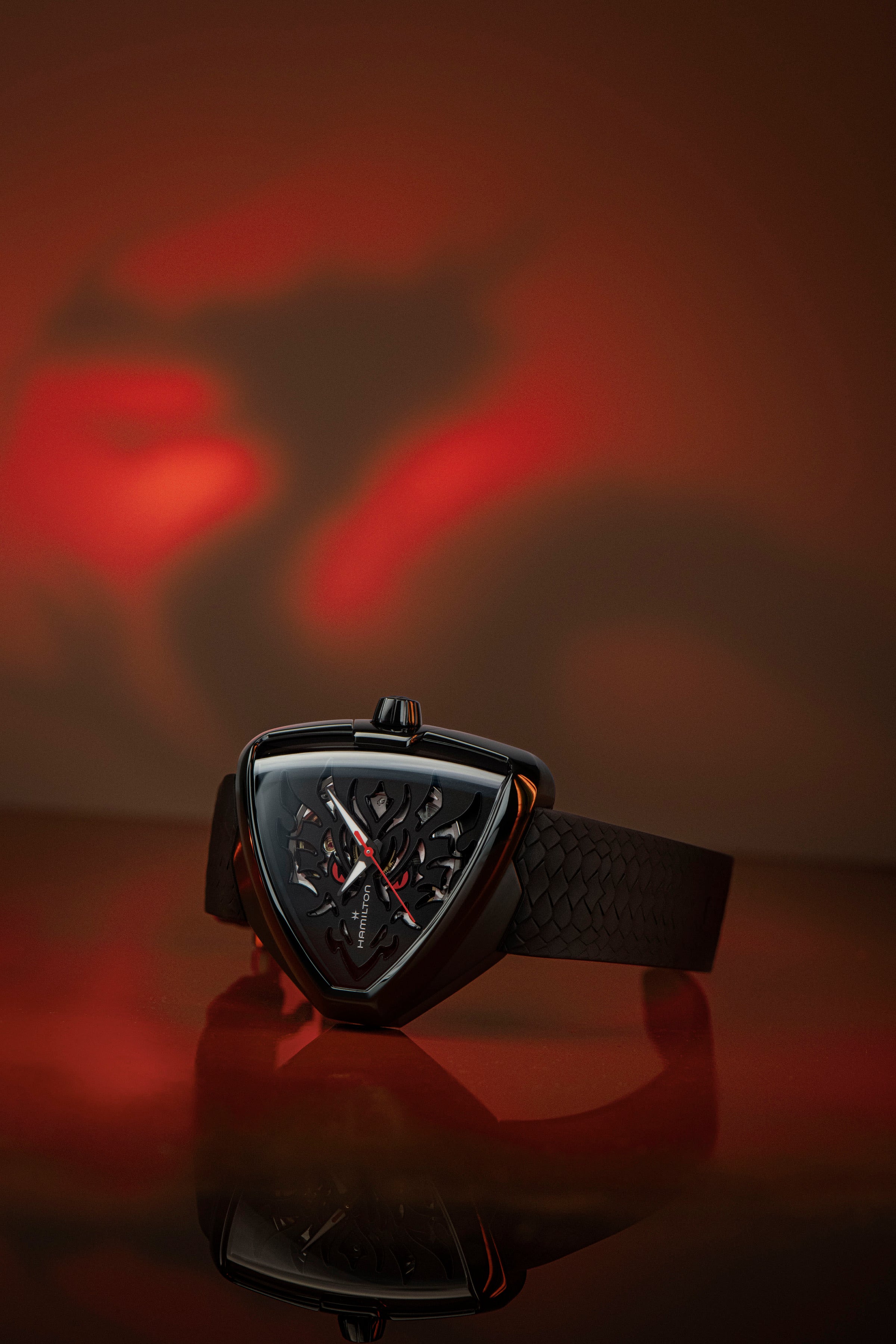 A Hamilton watch laid on its side against a red and black background. The watch features a triangular black dial and black strap.