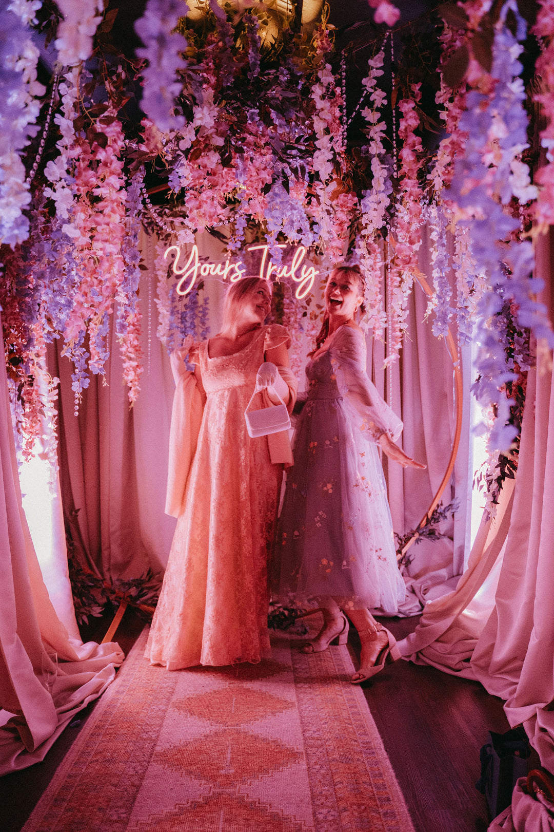 Two girls wearing gowns posing in front of a Yours Truly backdrop with floral decorations.