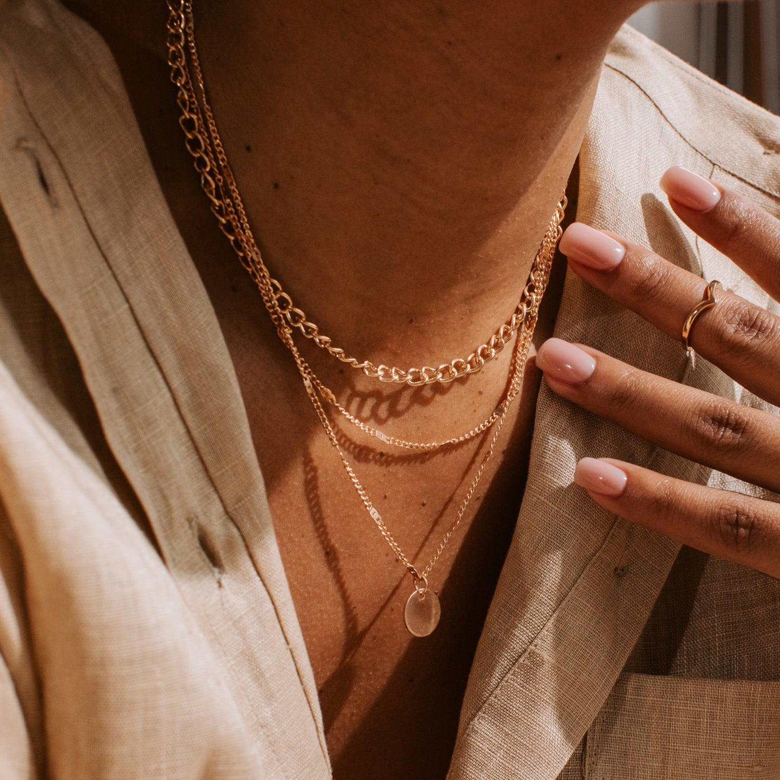 photo of 3 gold necklaces hanging on a woman’s chest, wearing a tan button up shirt, with her hand resting on her lapel wearing a gold ring on her first knuckle