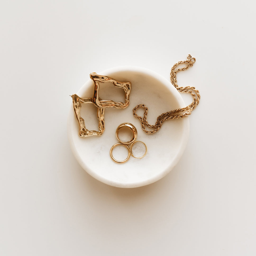 photo of a white jewelry dish with a pair of gold earrings, a gold necklace and 3 gold rings, sitting inside the dish