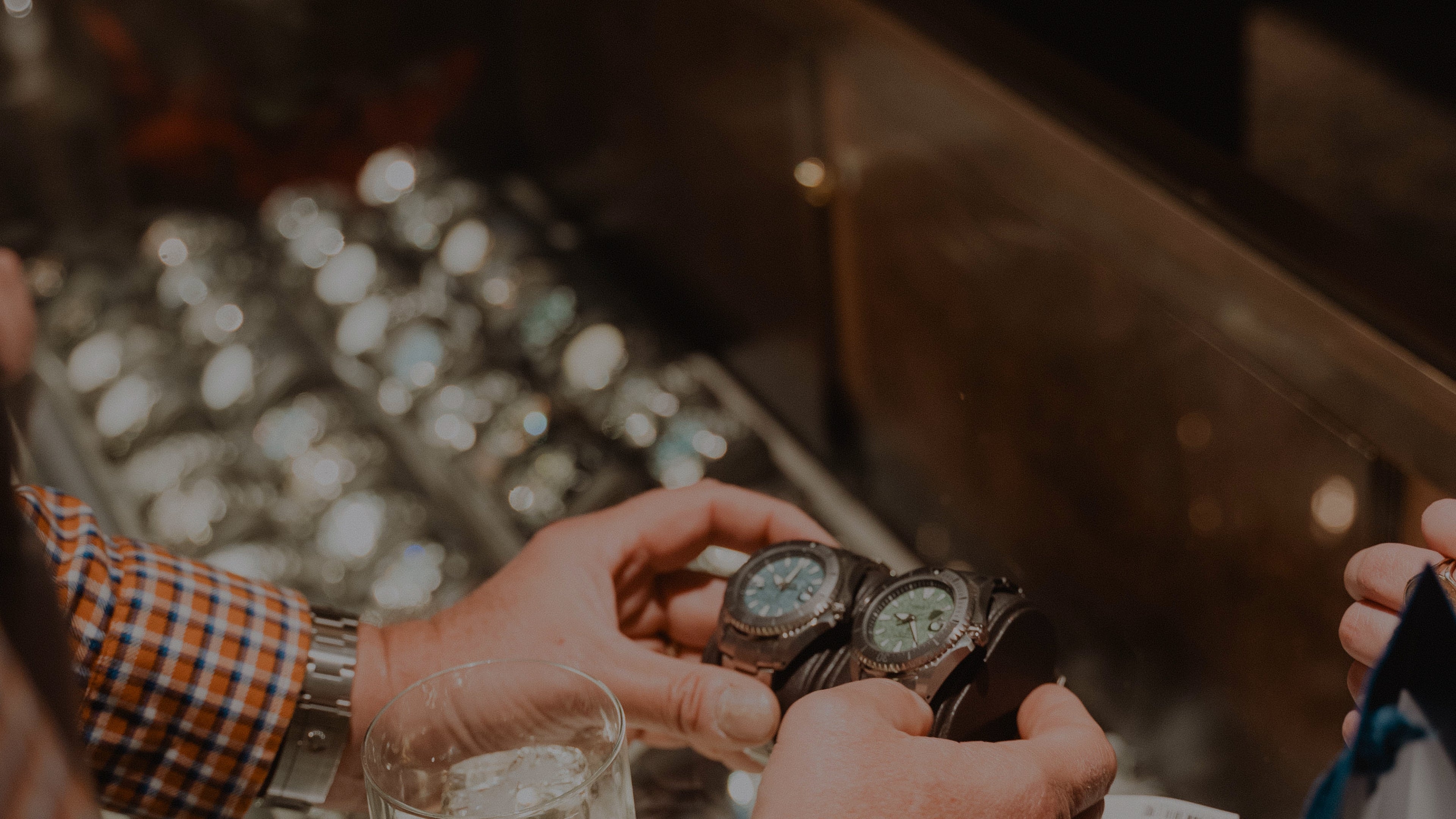 A close-up of a man holding two watches beside each other over a display glass. The two watches feature a green and a blue dial.