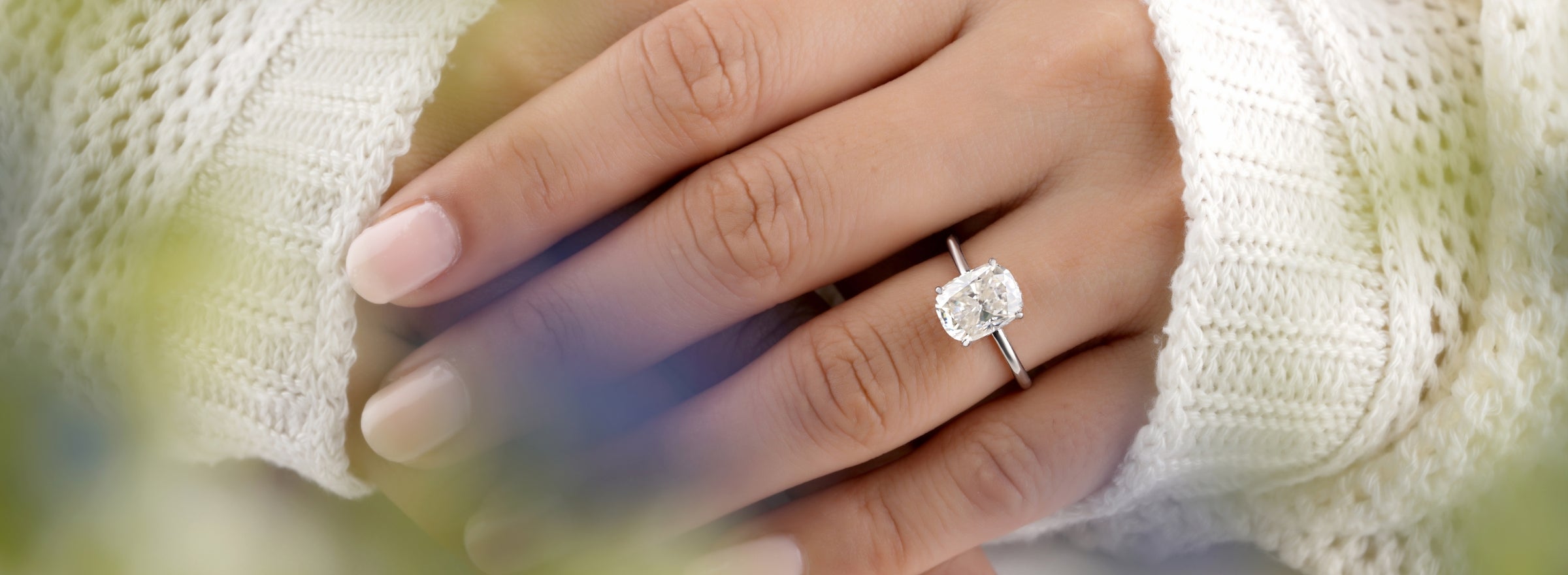 This is a close-up of a woman's hand with light pink nail polish. She is wearing a white sweater and showcasing her diamond engagement ring.