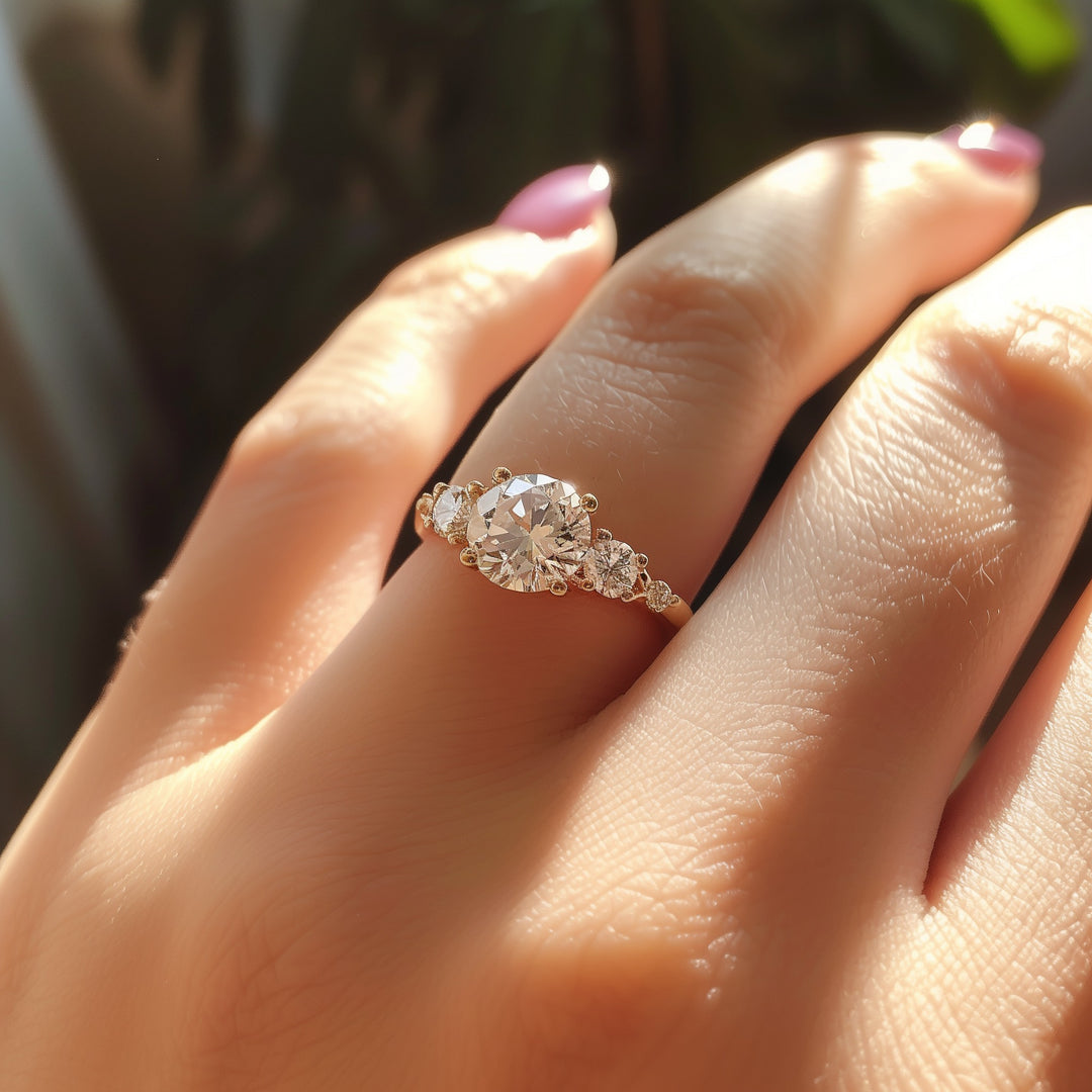 A close-up of a woman's hand showcasing her yellow gold engagement ring on her wedding finger. The ring features a round-shaped center stone and side stone diamonds set with prongs.