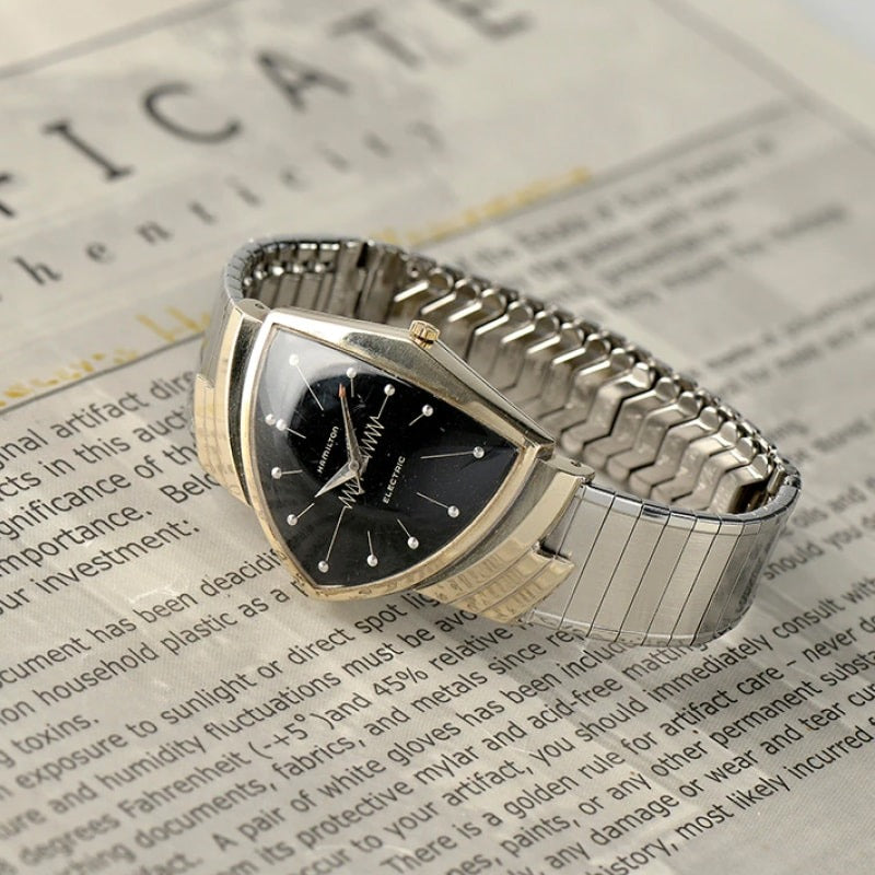 A Hamilton watch is laid on its side against a newspaper. The watch features a triangluar black dial, stainless steel case, and bracelet.