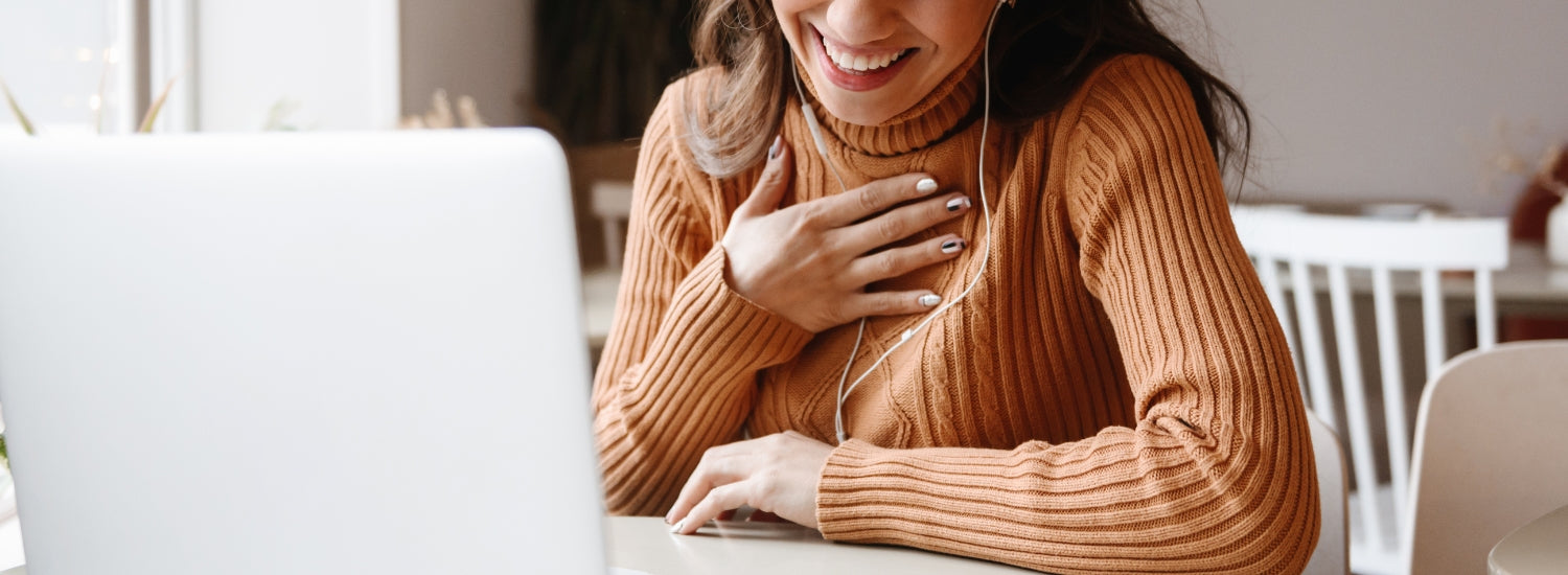 photo of a woman in a light orange corded sweater holding her hand to her chest while she smiles looking down at her laptop screen