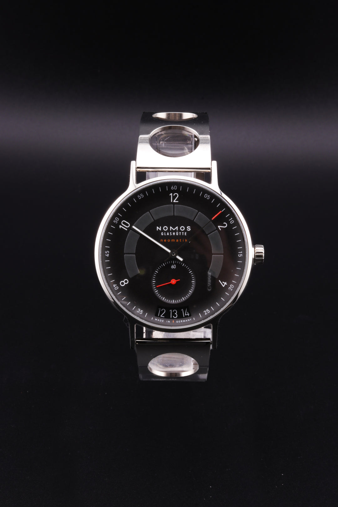 A NOMOS watch is displayed in the middle of a gray background. The watch features a black dial, a circular stainless steel case, and a stainless steel bracelet with open-circle designs.