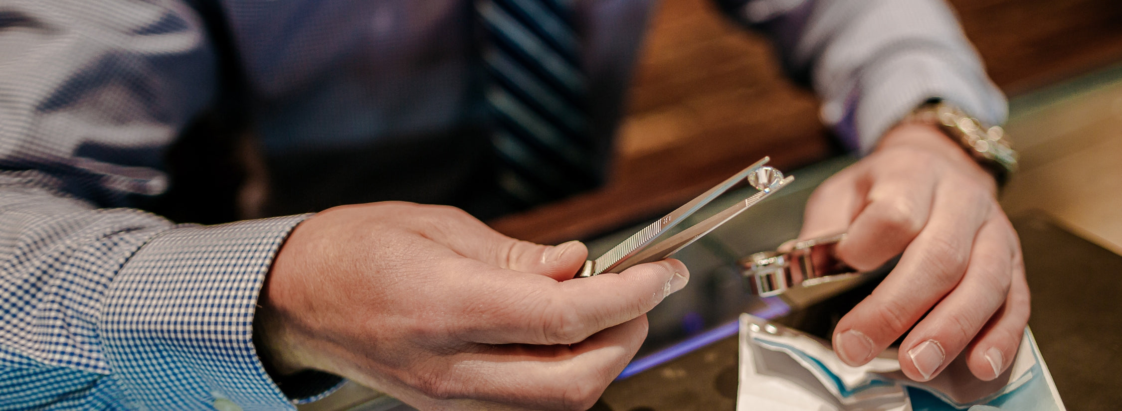 photo of a hand in a suit holding a loose diamond in tweezers while holding a magnifying glass in the other hand