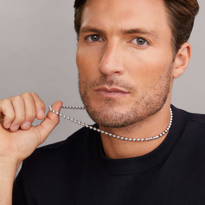 A close-up of a man with short hair and facial hair wearing a black shirt and holding his white gold ball chain necklace by his thumb.