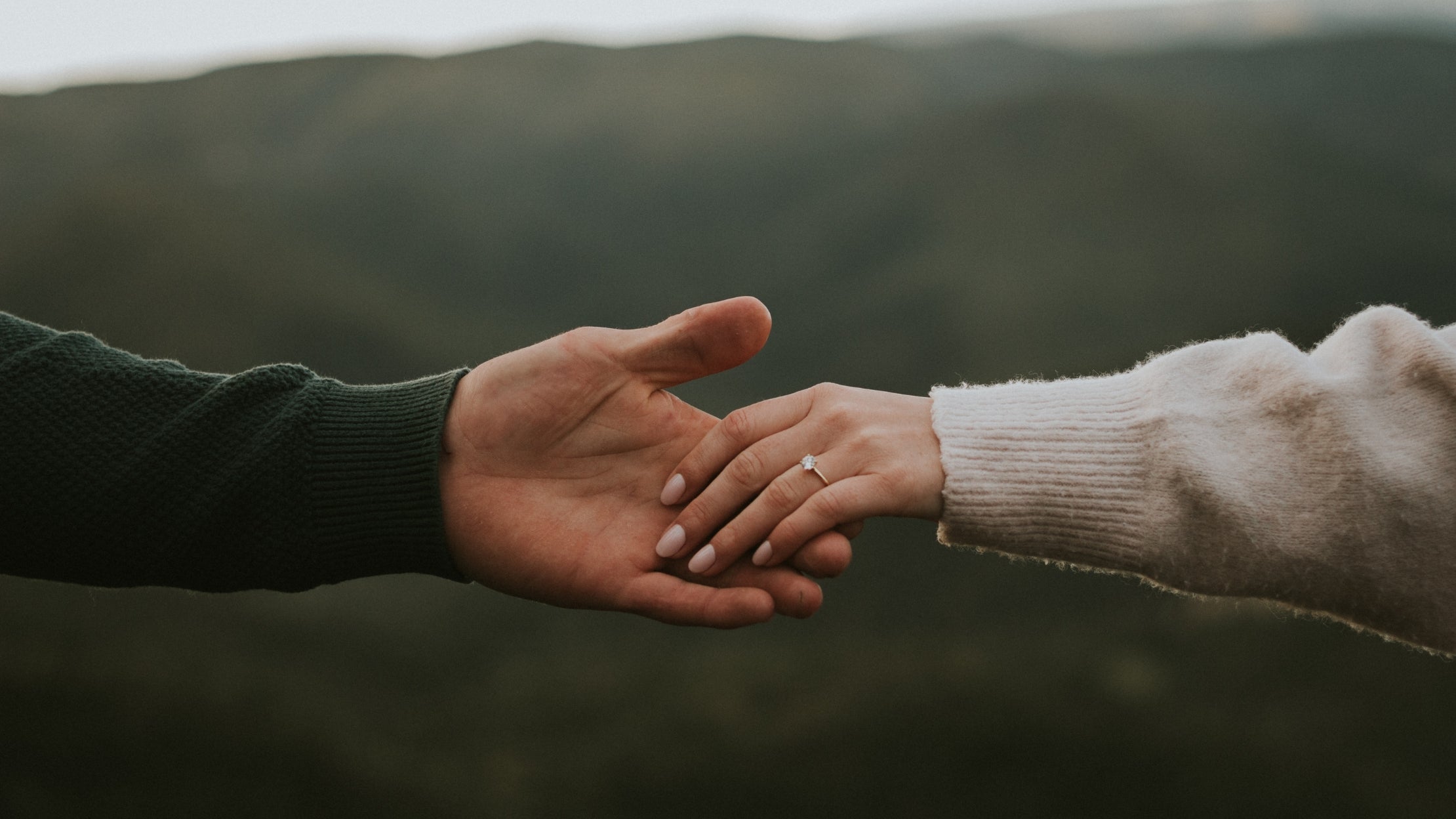 photo of a mans hand reaching out and a woman’s hand, wearing a diamond engagement ring