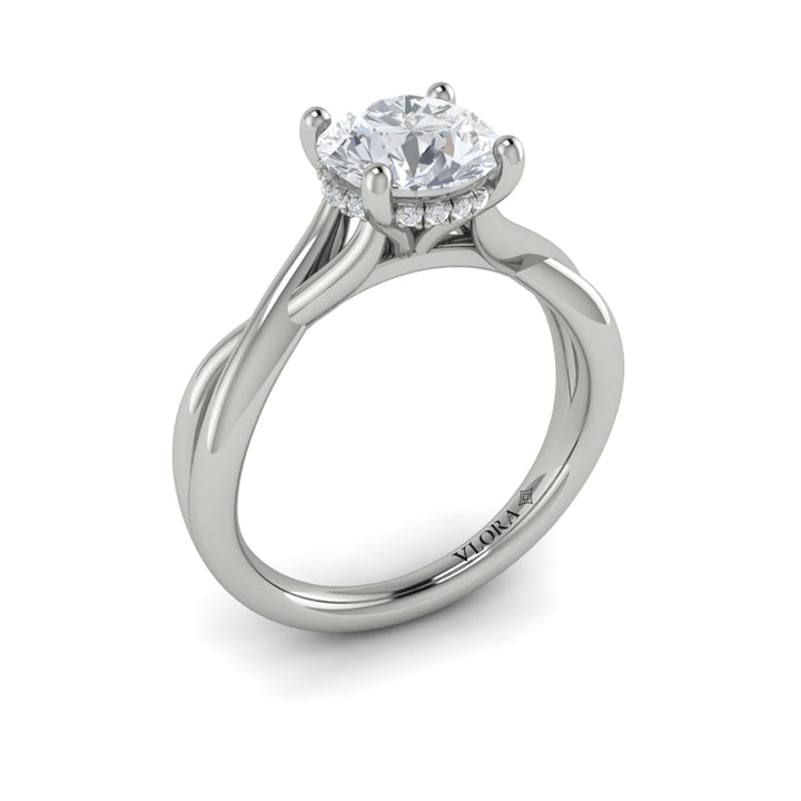 An angled view of a white gold engagement ring is set against a white background. The engagement ring features a round center diamond, a cathedral setting, a hidden halo, and a twisted band design.