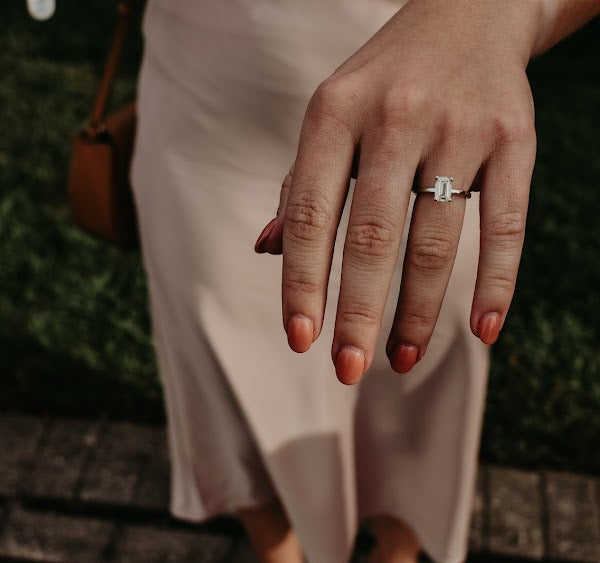 A woman is holding her hand out showing her engagement ring. The ring features an emerald-shaped center stone and a yellow-gold band.
