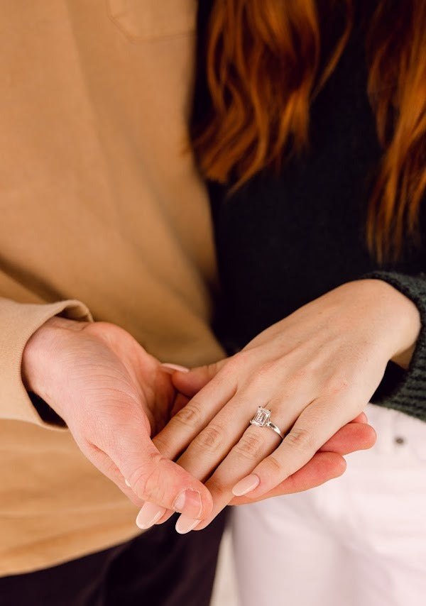 A close up of a couple holding hands showcasing her engagement ring. The ring features an emerald-cut center stone and a white gold band.