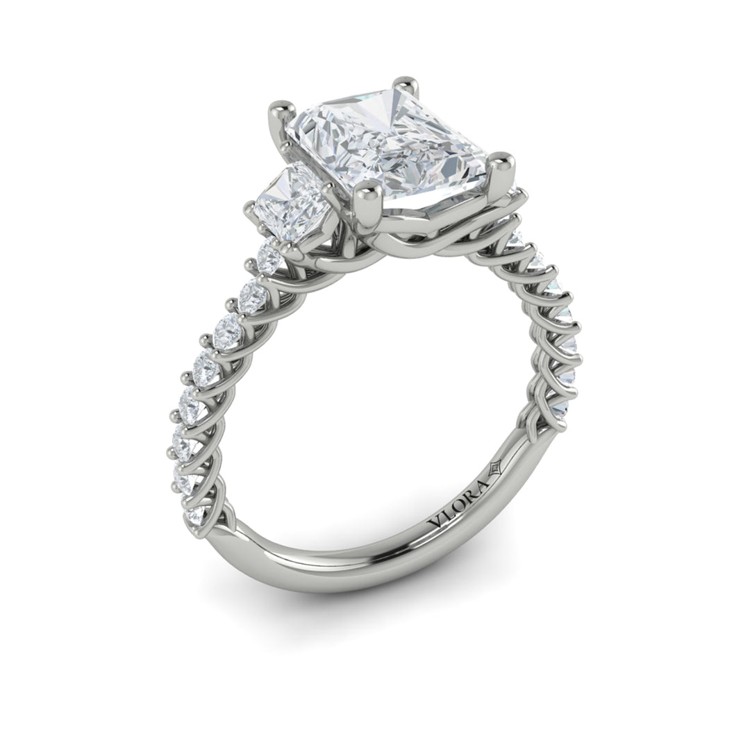 An angled view of a white gold engagement ring is set against a white background. The engagement ring features a center emerald cut diamond flanked by two emerald cut side diamonds, an intricate woven setting, and a diamond-set band.