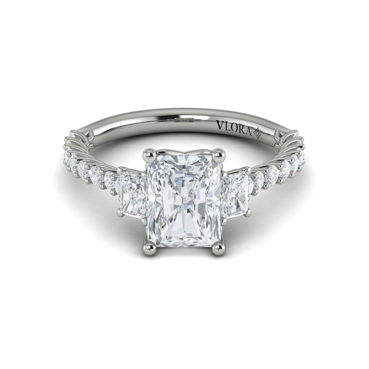 A front view of a white gold engagement ring is set against a white background. The engagement ring features a center emerald cut diamond flanked by two emerald cut side diamonds, an intricate woven setting, and a diamond-set band.