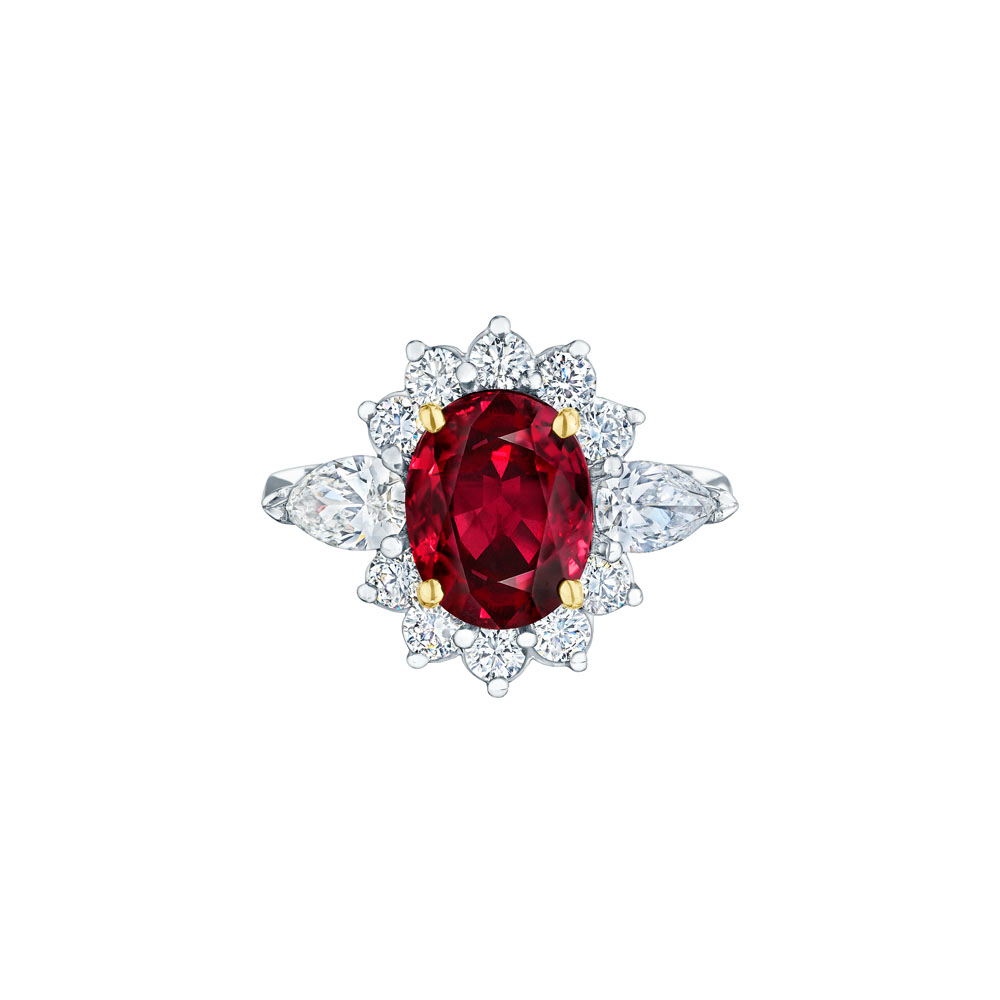 A white gold ring is in the middle of a white background. The ring features an oval-shaped ruby center stone set with yellow gold prongs with round and marquise-shaped side stone diamonds.