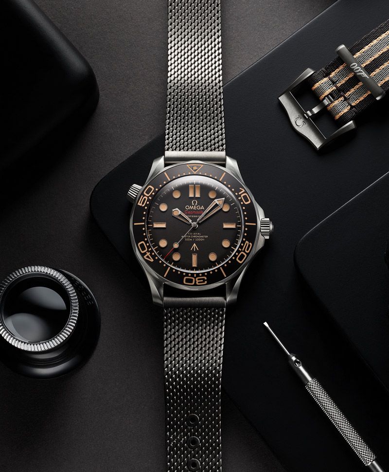 An OMEGA Seamaster watch laid out against a gray background with other watch accessories. The watch features a black dial, tan hands and markers, a black bezel, and stainless steel case and bracelet.
