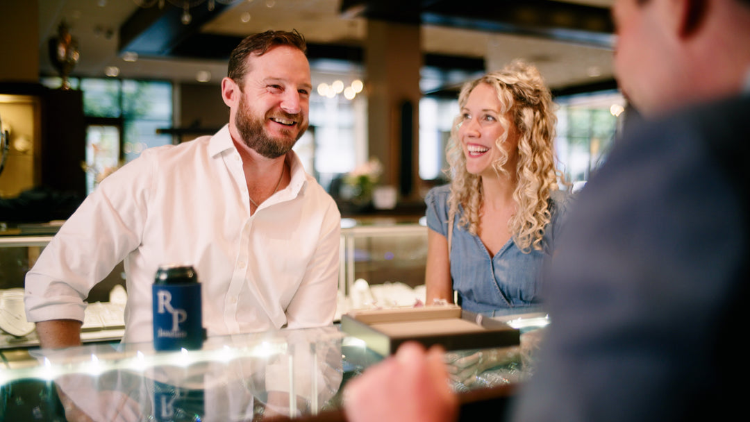 photo of a brunette man with short hair and a beard wearing a white button down shirt sitting next to a smiling blonde curly haired woman wearing a chambray dress - sitting at a glass counter top across from a man