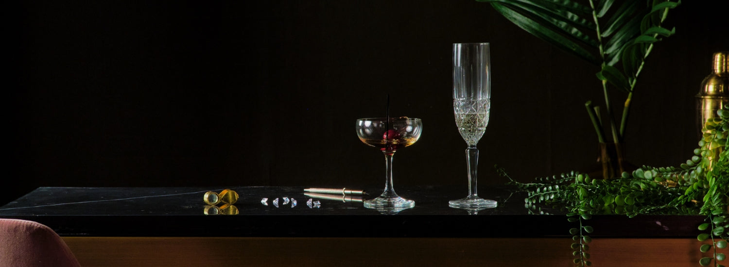 photo of a gold object sitting next to several loos diamonds, a coup glass with a cherry in it, a champagne glass, greenery and s gold shaker