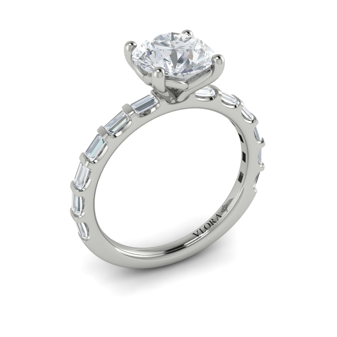 An angled view of a white gold engagement ring is set against a white background. The engagement ring features a center round cut diamond, an Art Deco design, and a streamlined band of bar-set baguette diamonds.