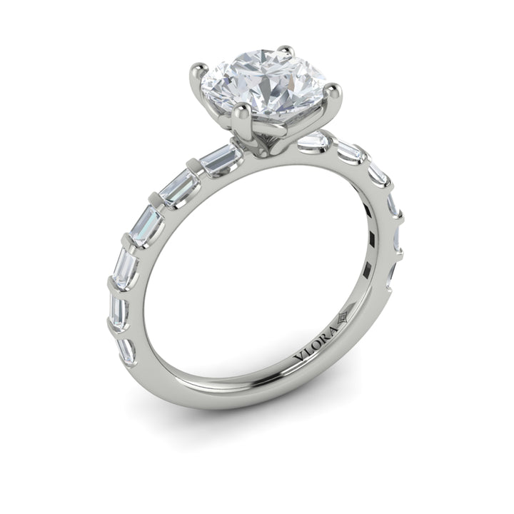 An angled view of a white gold engagement ring is set against a white background. The engagement ring features a center round cut diamond, an Art Deco design, and a streamlined band of bar-set baguette diamonds.
