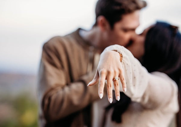 A man and woman are kissing, blurred in the background, and focusing on the woman's marquise-shaped engagement ring on her wedding finger.