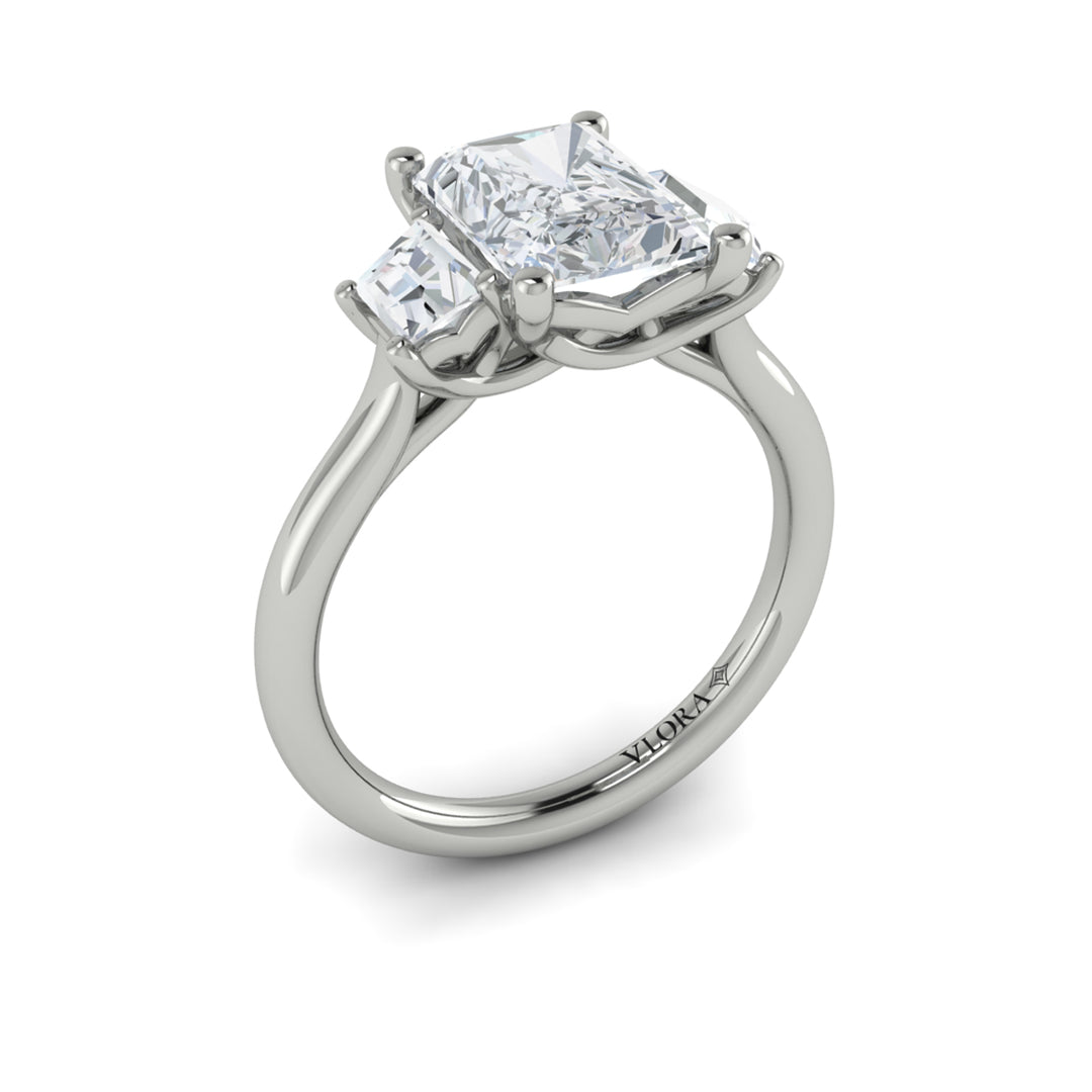 An angled view of a white gold engagement ring is set against a white background. The engagement ring features an emerald cut center diamond, flanked by complementary emerald cut side stones, in a woven setting.