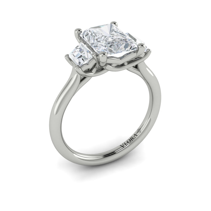 An angled view of a white gold engagement ring is set against a white background. The engagement ring features an emerald cut center diamond, flanked by complementary emerald cut side stones, in a woven setting.
