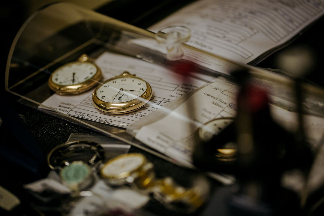 A black cluttered desk with a case over three stopwatches and paperwork, one wristwatch, and more paperwork in the background.