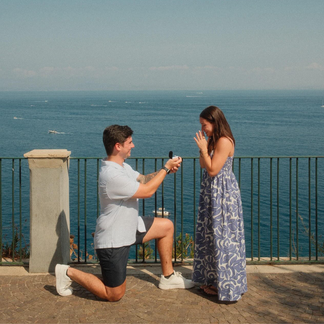 A close up of a man on his knee proposing to a woman in front of the ocean.