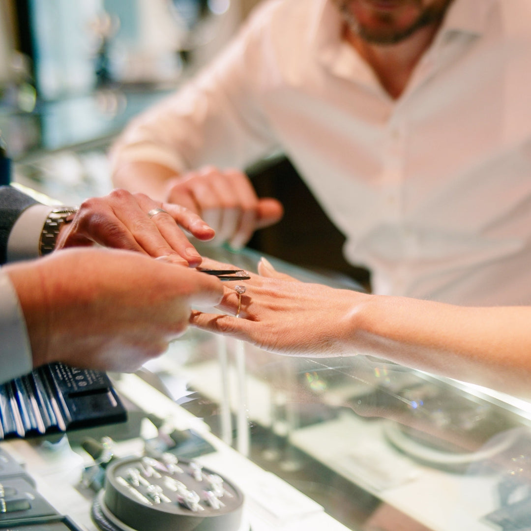 A close-up of a woman holding out her hand with her engagement ring on her finger, with her partner wearing a white button-down next to her. She is reaching out to a jeweler to place a ring on the band.