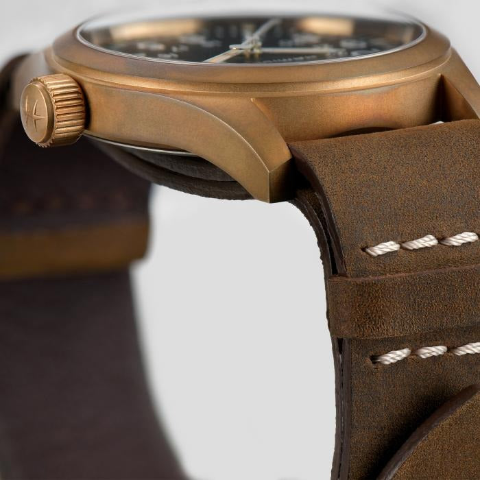 A close-up side-view of a Hamilton watch, focusing on the dial, bronze case, a bronze crown, and a brown leather strap.