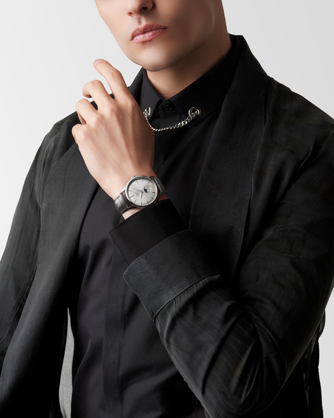 A close-up of a man holding his hand to his chin with a watch around his wrist. The watch features a silver dial, silver hands and markers, a stainless steel bezel, a crown on the right side, and a black leather strap.