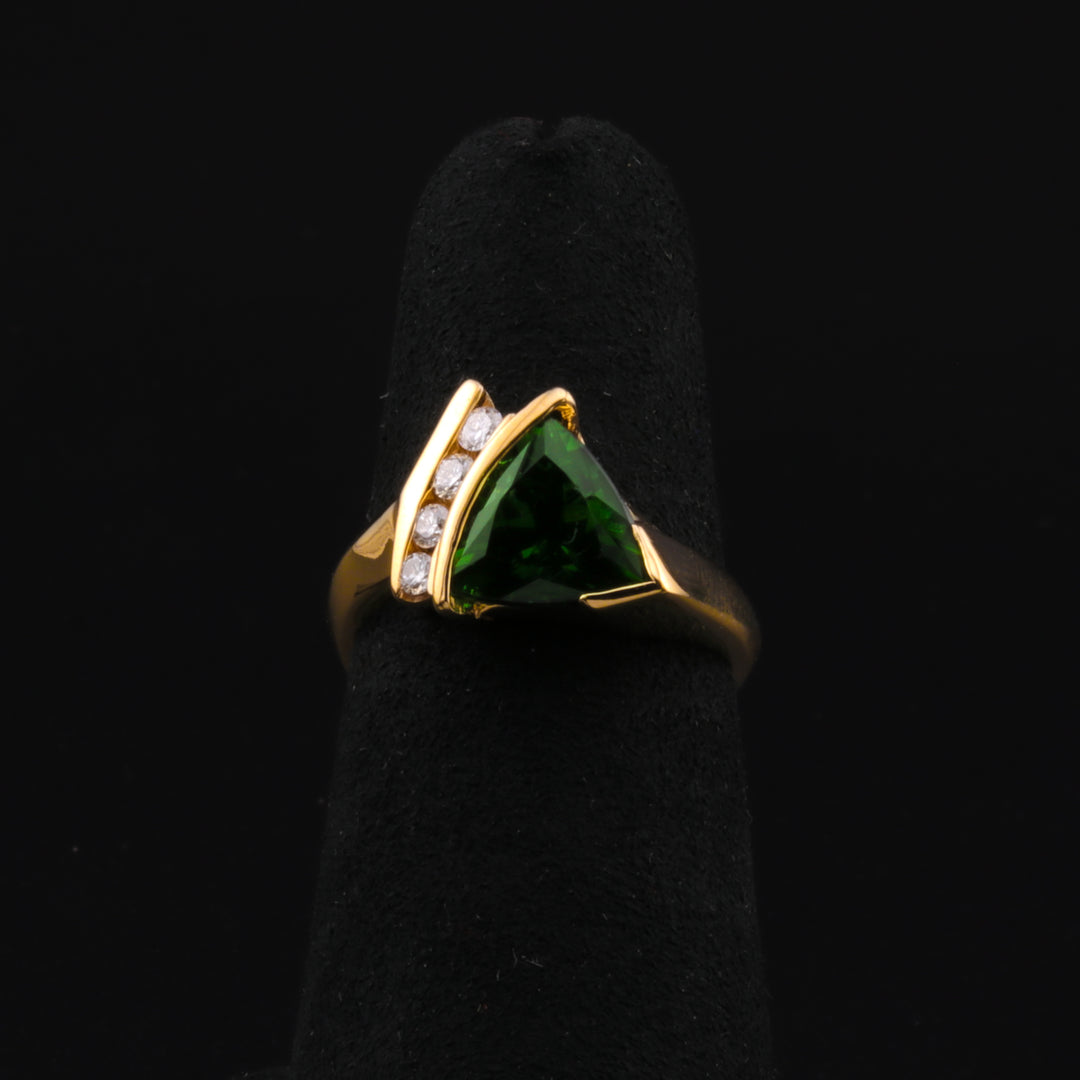 A close-up of a yellow-gold neo-expressionist ring with a trillion-cut green tourmaline and four sparkling diamonds, set on a luxurious yellow gold band resting on a black ring holder against a black background. 