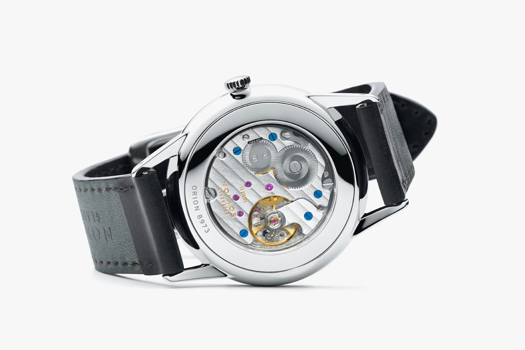 The back of a Nomos Glashutte watch rests on the right side, showcasing the back of the dial, stainless steel bezel, and strap.