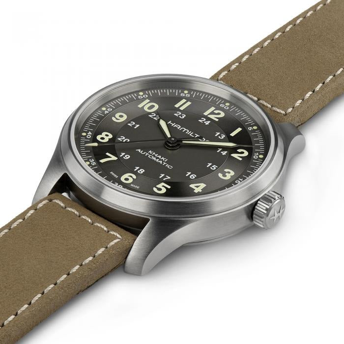 A Hamilton watch is laid out tilted to the right against a white background. It features a black dial, light green hands and markers, a stainless steel case, bezel, and crown, and a brown calf leather strap.