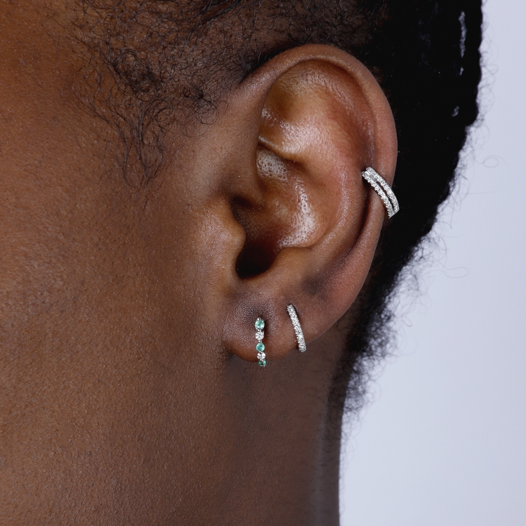 A woman with short black hair is wearing a pair of huggie earrings with alternating green and white diamonds. 