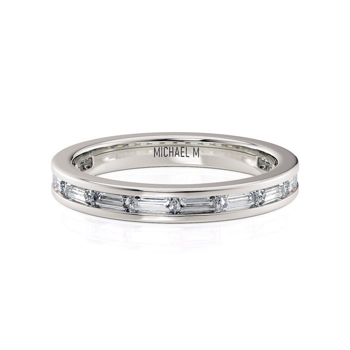 A white gold ring lies flat against a white background. It features a diamond set band with princess-cut diamonds, and the "Michael M" inscription is visible inside it.