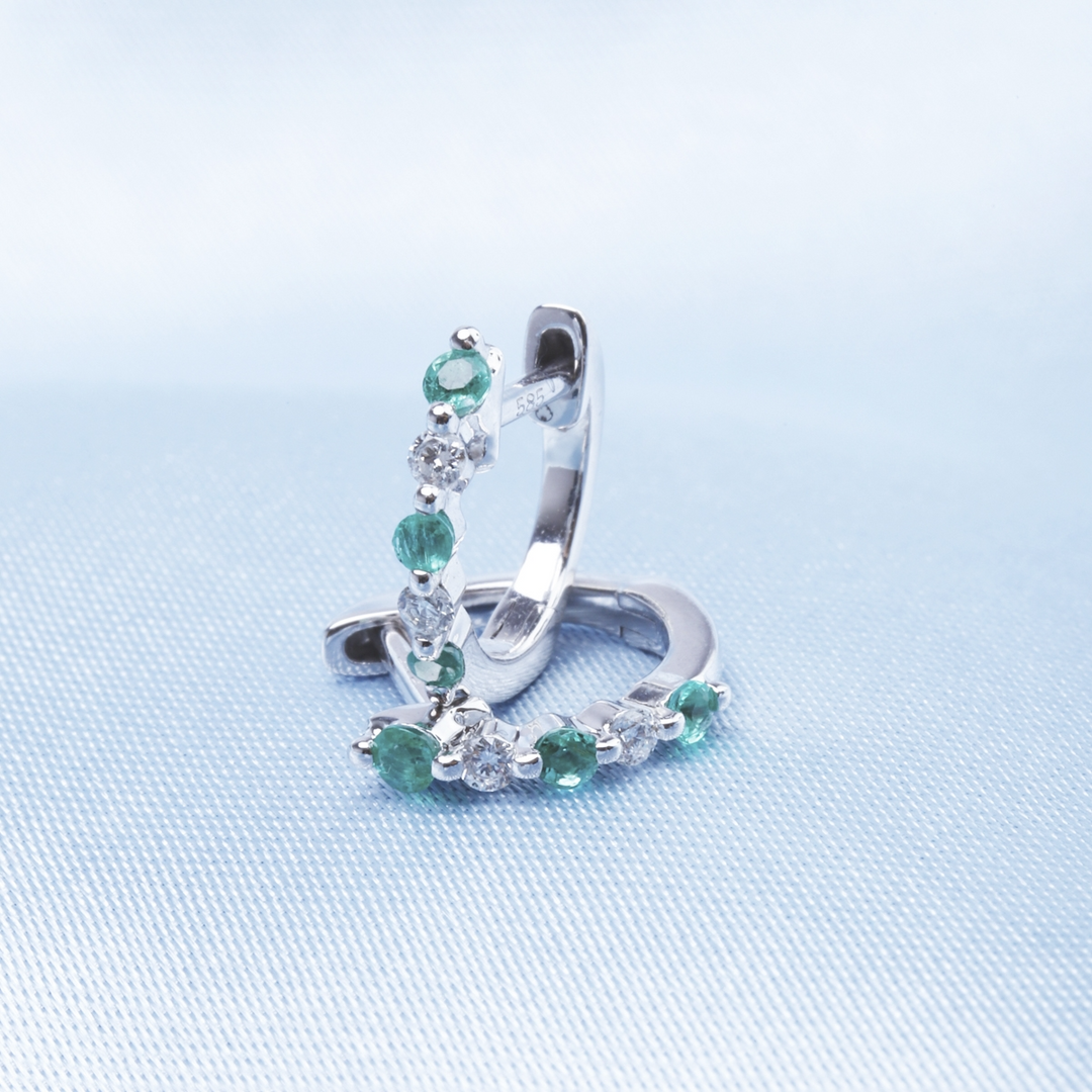 A pair of huggie earrings with alternating green and white diamonds. One earring rests on the light-blue surface, and the other is standing up.