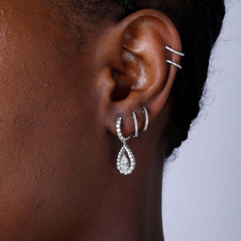 A woman with short black hair is wearing a pair of diamond dangle earrings with a teardrop shaped diamond in the center with a diamond set halo.