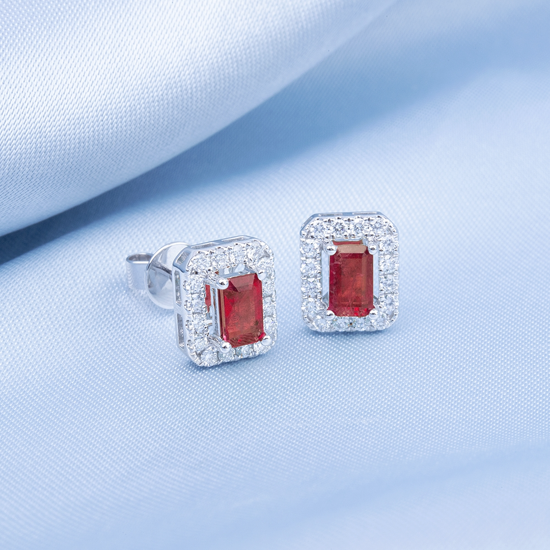 A pair of stud earrings, a ruby emerald-shaped diamond in the middle, and a surrounding diamond frame lied against a light-blue background. 